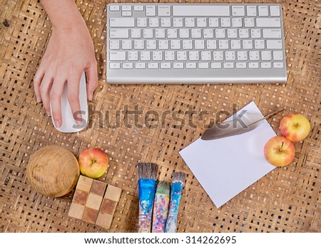 office wooden table with woman hand on computer mouse with computer keyboard with  paint brush, paper,  and apple , top view