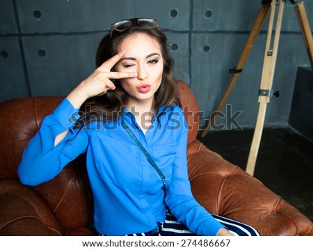 Young happy woman winking and showing tongue.Young student reading a book and sit on black office chair and showing victory by finger,Wear blue shirt,glamour glasses and  having fun.Office style