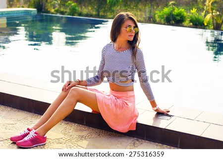 Fashion portrait of young sensual brunette woman with long amazing hairs, natural make up.Amazing and sunshine model in summer outfit sitting near pool in round sunglasses.