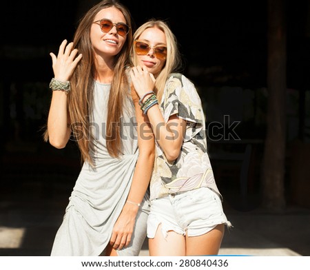 Two young brunette and blonde best friends looking on camera and laughing, have sexy slim body, wearing stylish dresses,sunglasses and fashion bright jewelry, posing in front of shadow.not isolated.