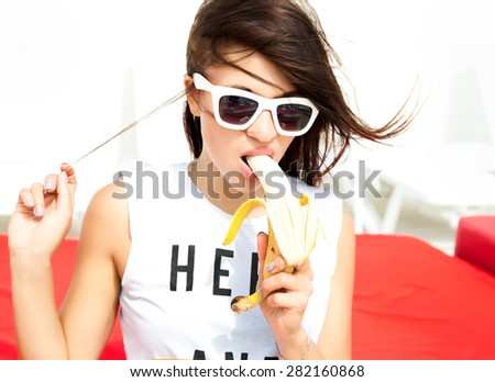 Young pretty teen girl with wearing stylish hipster outfit, and eating banana.\
Party concept.posing on the beach resort background, bright summer fashion portrait.