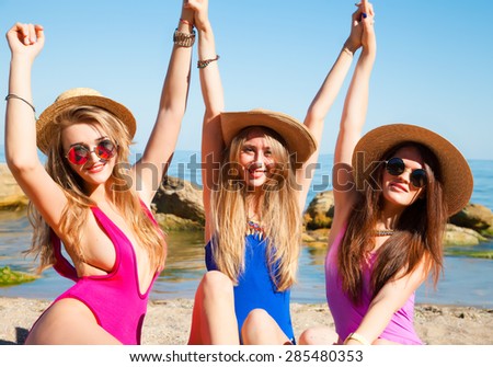 Three beautiful young women in bikinis dancing backlit on a sunny beach,friendship,put friends put their hands up,sunbathing on the beach.stylish girls in bathing suits and neon reflective sunglasses
