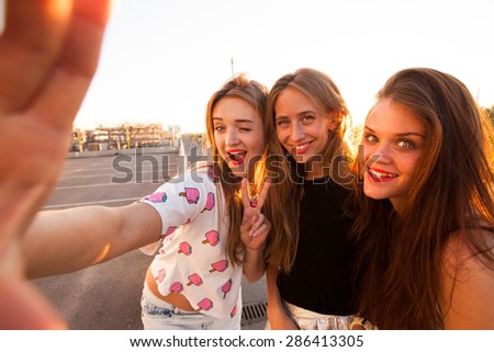 Close up fashion lifestyle portrait of tree young hipster girls best friends, wearing bright make up and similar trendy outfit,making funny faces and have gait time.Urban background.girls make selfie