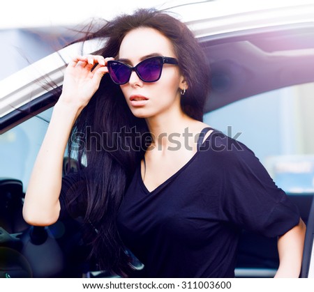 Portrait of beautiful sexy fashion woman model in summer hat and in black dress and luxury accessories with bright makeup sitting in luxury car. Young woman driving on road trip on sunny summer day.