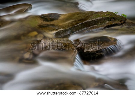 Water rushing by a rock in a river forming a smooth, abstract, painted appearing pattern.