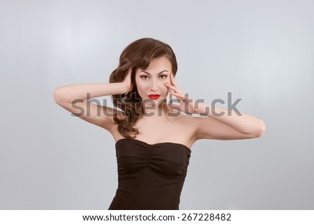 the beautiful young girl with dark hair with red lips and in a dark dress on a light background costs, one hand is held in hair, by the second on a cheek