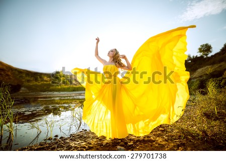 the beautiful girl with long hair in the yellow fluttering dress costs on the bank of a stream, hands raised up, having closed eyes