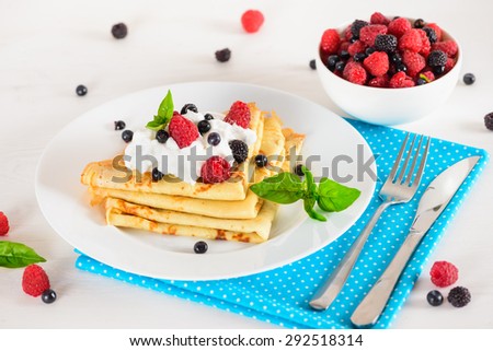 Delicious sweet crepes decorated air cream and ripe berries, raspberries, blackberries and blueberries on a white plate, fork, knife, bright blue polka dot napkin. Tasty breakfast .