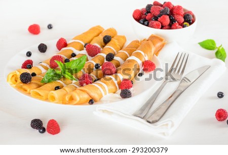 Delicious sweet crepes rolled in a tubes decorated cream and ripe berries, raspberries, blackberries and blueberries on a white plate, fork, knife, white napkin. Tasty breakfast .