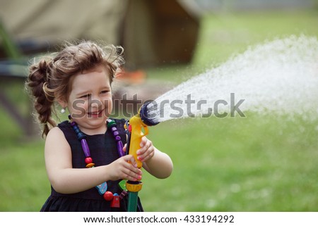 Pretty little girl dressed in a black dress and red boots watering grass