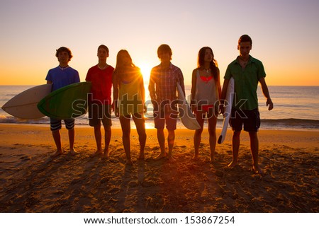Surfers teen boys and girls group walking on beach at sunshine sunset back light