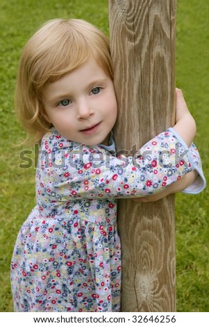 Beautiful little toddler blond girl hug a trunk with grass park background