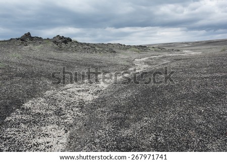 Small white dry river crosses a volcanic soil made by small black stones and disappears over the horizon coinciding with a gray cloudy sky