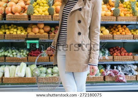 Girl in a beige coat and light blue jeans in the grocery store holding a shopping basket. In the background you can see the showcase of vegetables and fruits.