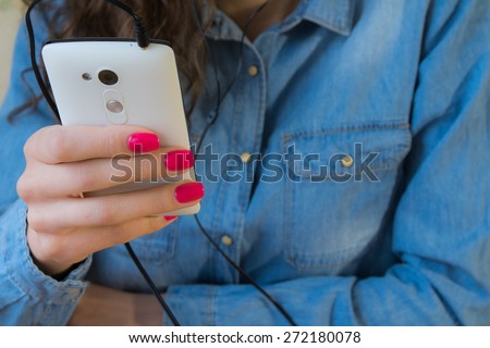 The girl in blue denim shirt with red nail Polish listens to music on a white phone.