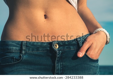 Belly of a young slim girl close-up. Girl dressed in blue jeans, her hand white clock. In the background the sea. Retro colors.