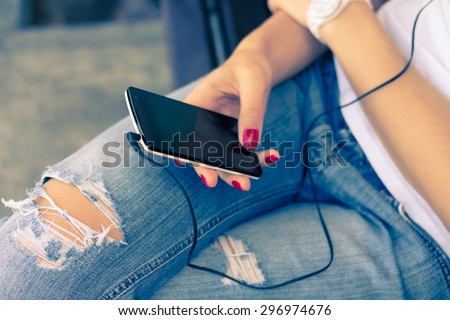A young woman in jeans sitting on the bench and connect the headphones to her mobile phone. White smart phone is in female hands with a red manicure. Listening to music while walking. Retro colors.