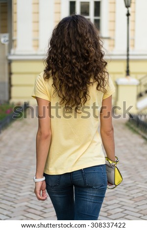 Attractive slim young woman with curly brown hair is walking on the old European city. She is wearing a yellow T-shirt and blue jeans, ladies handbag in her hand.