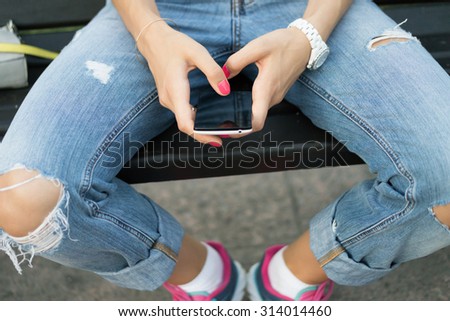 Female hand holding a mobile phone close-up. The girl in jeans and sneakers sitting on a city bench.