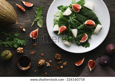 A salad with green leaves of rocket and spinach, fig pieces and melon in a white plate on dark wooden background. Top view.