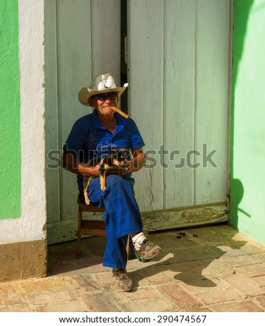 TRINIDAD, CUBA -DECEMBER 24, 2013: An unidentified old Cuban man sitting in a chair in front of the building in old part of Trinidad, Cuba with a cigar in his mouth and a dog on his lap.