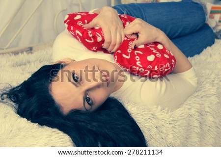 Beautiful girl lying down and hugging a cushion. Pillow is red as a heart. The girl looks in the picture, the face in focus and close-up. The girl\'s dark hair  are scattered on the bedspread.