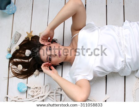 Beautiful girl lying on the floor. Brunette girl , in her hair are shells . She is wearing a white dress . She holds hands near the face . Lovely girl like a mermaid. Portrait . Studio