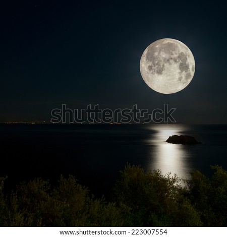 Full Moon and moonlight on night sea. City lights on the horizon.