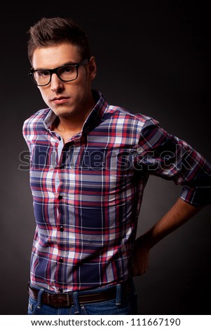 Young casual man in a squared shirt and eyeglasses is holding his jeans at the back and is looking seriously at the camera. over black background