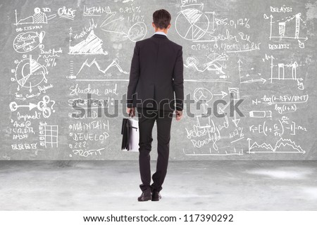 back view of a business man holding a briefcase and looking at some charts , graphs and calculations on a blackboard