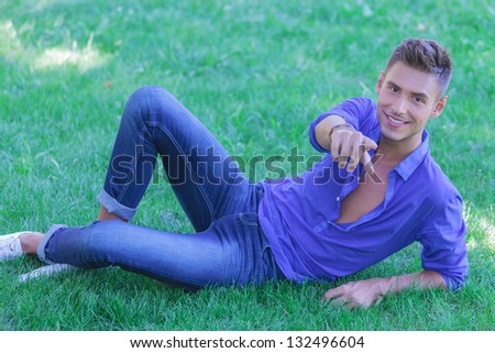 young casual man laying in the green grass and pointing at the camera while smiling