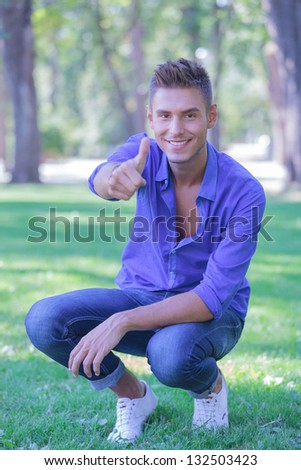young casual man in a crouched position in the park is showing thumb up gesture and smiling to the camera