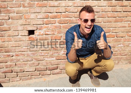casual young man sitting crouched by a brick wall and showing thumbs up with both his hands while smiling largely at the camera