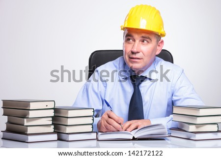 senior business man with helmet, sitting at a desk full of books and writing while contemplating, looking away from the camera. on gray background