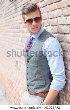 casual young man stands with his back against a brick wall with his hands in his pockets while looking away from the camera
