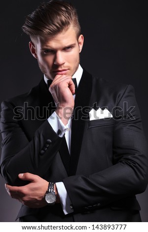 portrait of an elegant young fashion man in tuxedo looking at you with his hand at his chin and a pensive expression. on black background