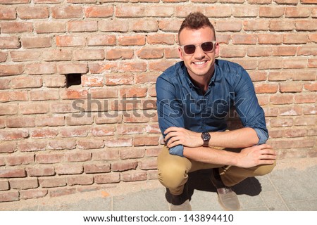 casual young man crouching in front of a brick wall while smiling for the camera