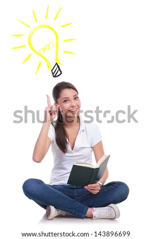 casual young woman sitting with legs crossed and having an idea while holding a book, pointing up at  a light bulb while looking at the camera. isolated on white background