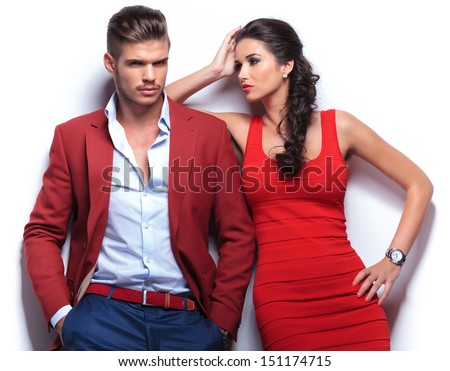 casual couple leaning against a white wall, man with hands in his pockets looking at the camera . woman looking at the man while leaning on his shoulder