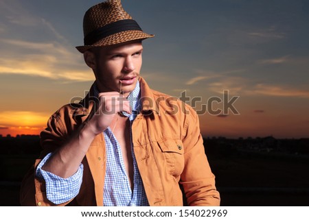 casual young man standing outdoor with the sunset behind him and holding a straw in his mouth while looking away from the camera