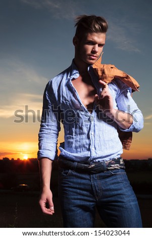 casual young man standing outdoor with his jacket over his shoulder with the sunset behind him