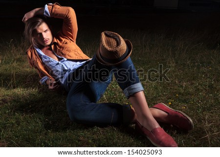 casual young man laying in the grass with his hat on his knee and his hand on his head while looking at the camera with a straw in his mouth