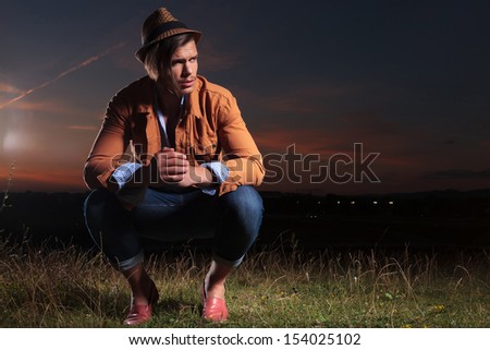 casual young man sitting crouched outdoor in the evening and looking away from the camera