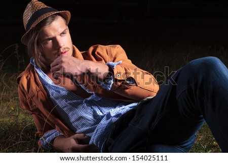 casual young man sitting outdoor and holding a straw in his mouth while looking away from the camera