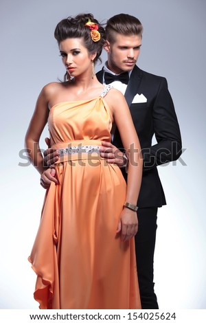 young fashion couple with man behind woman holding her by the waist while she looks at the camera. on a gray background