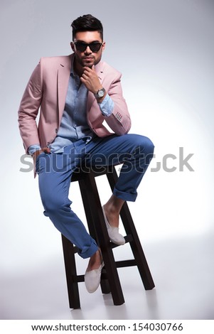 full length photo of a young casual man sitting on a chair and looking into the camera with his hand at his chin. on gray background