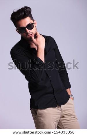 young fashion man sensually touching his lower lip while holding his other hand in his pocket and looking into the camera. isolated on a gray background