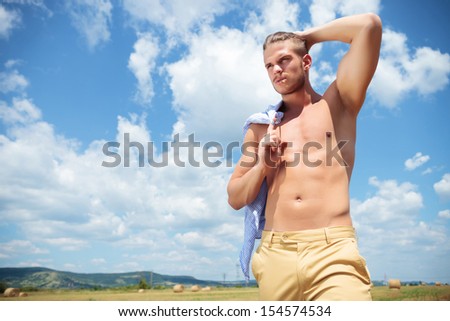 young topless man posing outdoor with his hand holding back his hair while holding his shirt on his shoulder and looking away from the camera