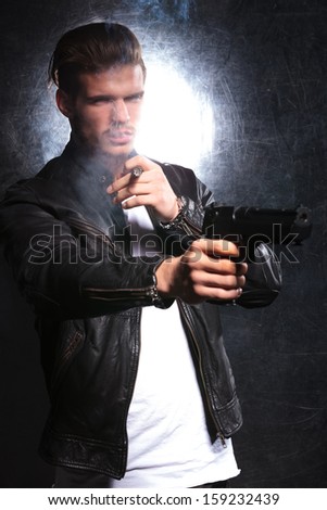 young fashion model dressed in leather jacket as a killer pointing his pistol away, while smoking a cigar