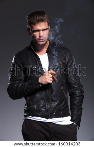 relexed man in leather jacket is smoking his cigar on a gray background